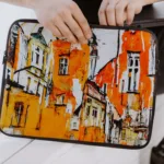 Aizsargājošs portatīvā datora maks tiek turēts rokās, dizainā abstrakti veidots vecpilsētas gleznojums oranžos toņos